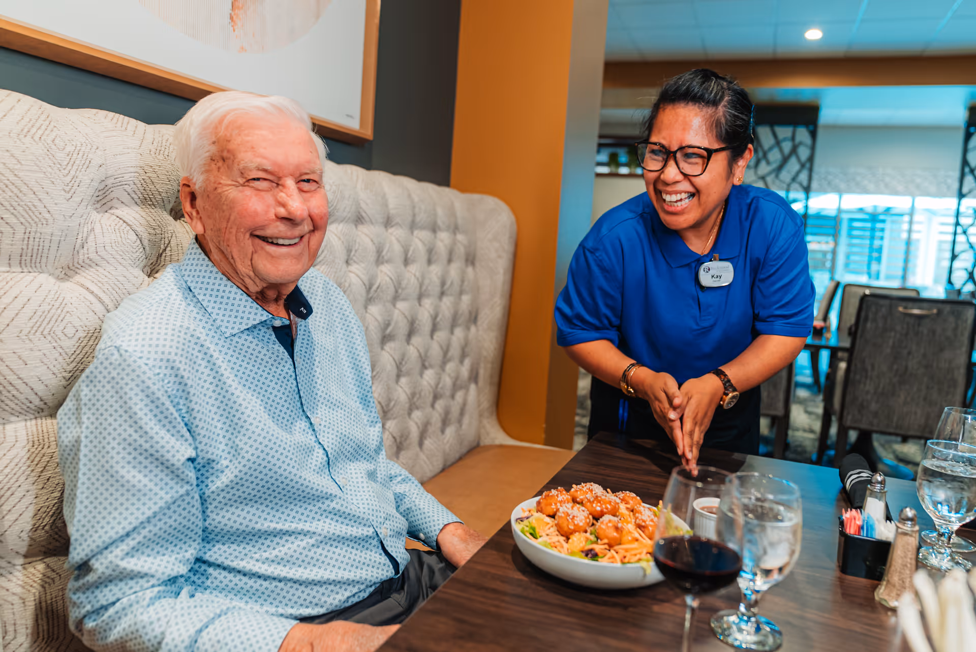 An elderly man sitting at a dining table smiling, while a woman in a blue shirt with a name tag labeled 'Kay' stands next to him, also smiling. On the table is a bowl of food, glasses of water, and a glass of red wine. The setting appears to be a dining area in a senior living facility.
