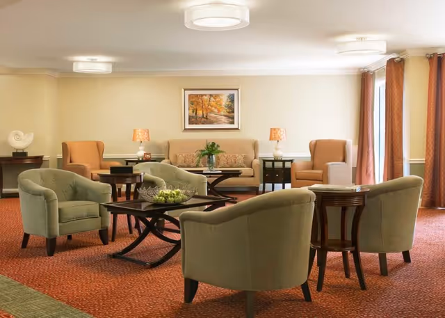A cozy living room with beige walls and red carpet featuring multiple armchairs and sofas arranged around dark wood coffee tables. There are two table lamps with orange shades on side tables, a framed autumn landscape painting on the wall, and large windows with orange curtains letting in natural light.