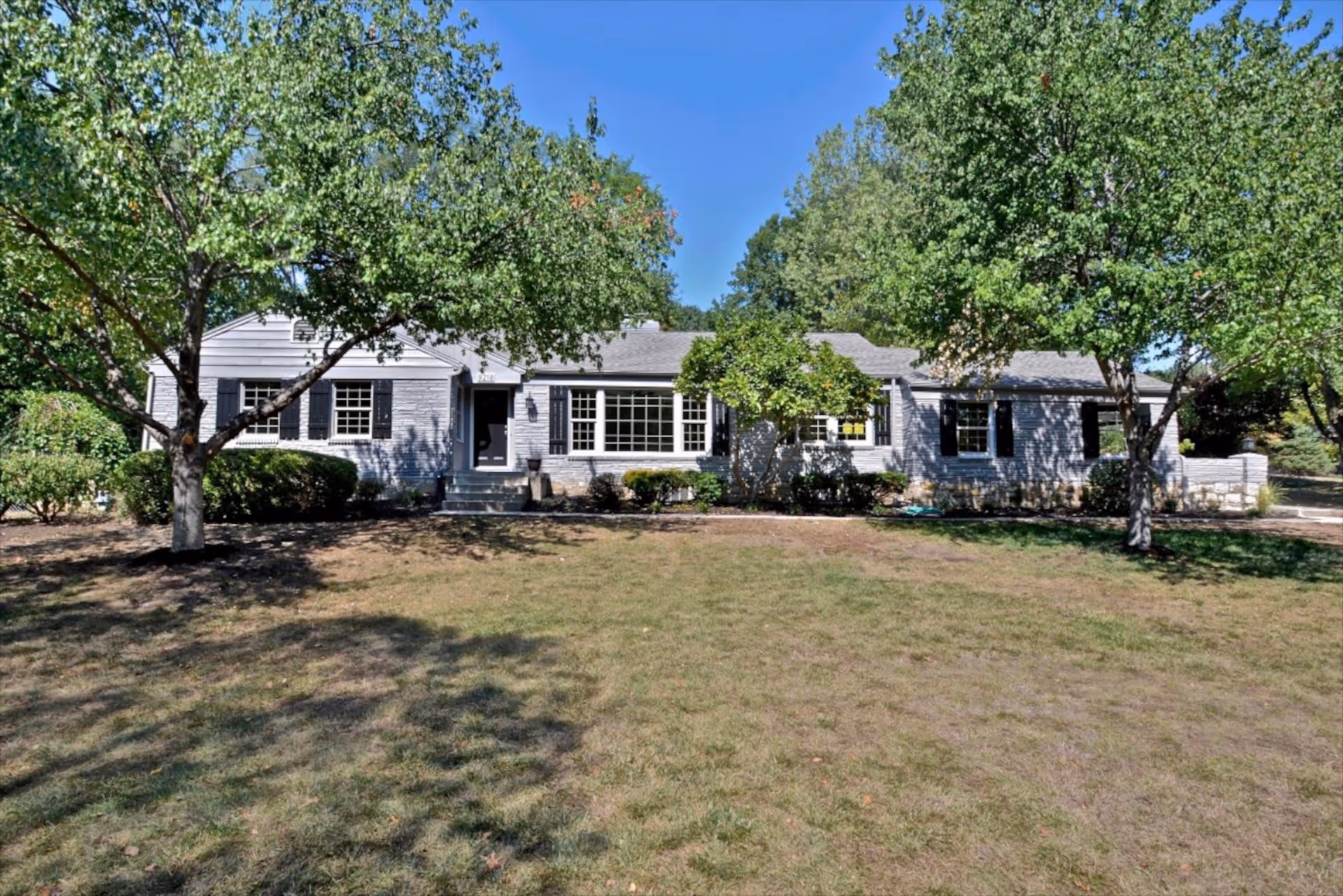 Front exterior view of a single-story residential building with gray siding, black shutters, and a central entrance door. The building is surrounded by green trees and bushes, with a grassy lawn in the foreground under a clear blue sky.