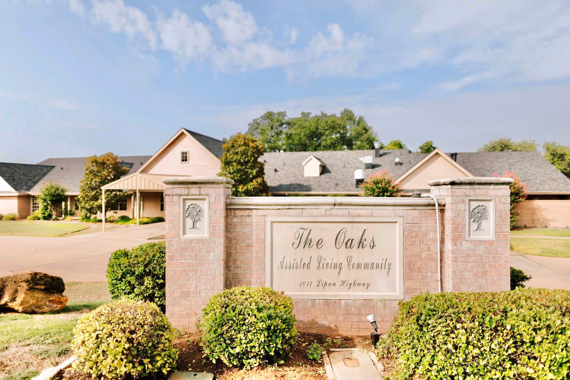 Brick entrance sign reading 'The Oaks Assisted Living Community' with shrubs and the facility buildings behind under a blue sky.