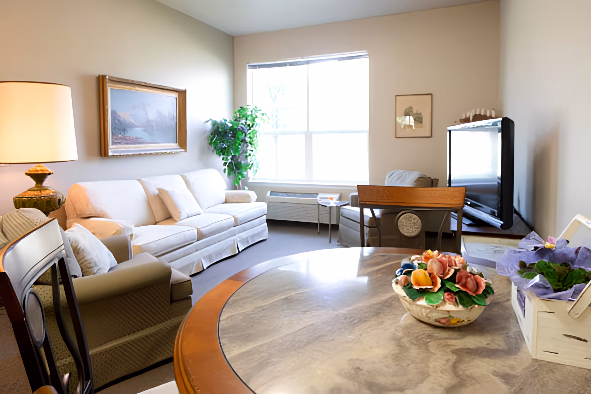A bright living room with a large window letting in natural light. The room features a white sofa with cushions, an armchair, a wooden dining table with a floral centerpiece, a lamp on a side table, a potted plant near the window, a framed painting on the wall, and a flat-screen TV on a stand.