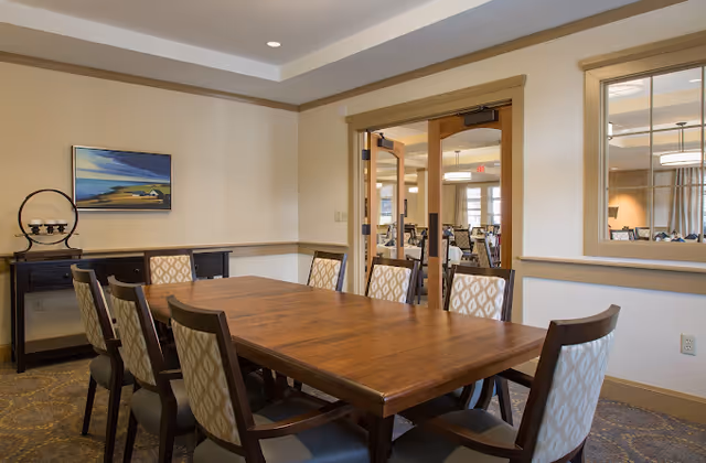 A dining room with a rectangular wooden table surrounded by eight upholstered chairs with patterned backs. The room has beige walls, a framed landscape painting, a small black sideboard with decorative items, and a large window opening into another room with more tables and chairs. The ceiling has recessed lighting.