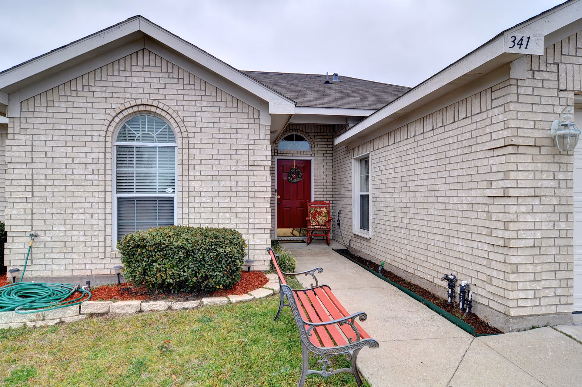 Front exterior view of a single-story brick residential building with a red door decorated with a wreath. There is a concrete walkway leading to the entrance, a red bench with black metal armrests on the right side of the walkway, and a green garden hose coiled on the left side near a bush. The house number 341 is visible on the right side of the roofline.