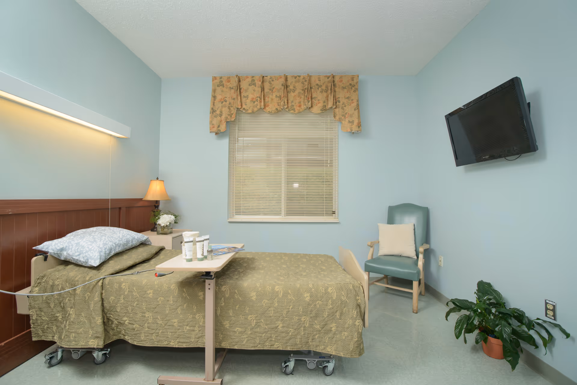 A small, simple bedroom with a hospital-style bed covered in a green patterned blanket and a blue pillow. There is a bedside table with a lamp and flowers, a window with floral valance and blinds, a green armchair with a white cushion, a wall-mounted TV, and a potted plant on the floor. The walls are painted light blue.