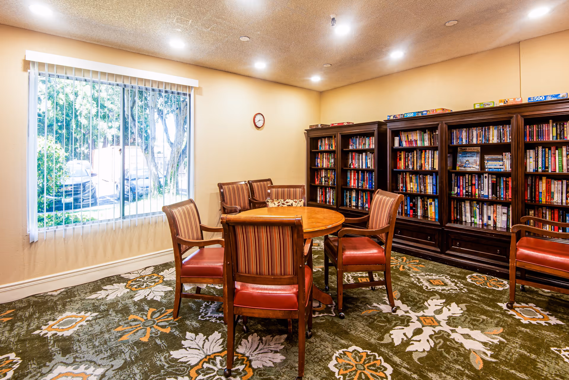 A cozy reading room with a round wooden table surrounded by six wooden chairs with red cushioned seats. The room has a large window with vertical blinds letting in natural light. Along one wall, there are three dark wooden bookshelves filled with books and puzzles on top. The carpet has a green, white, and orange floral pattern, and a small clock hangs on the beige wall.