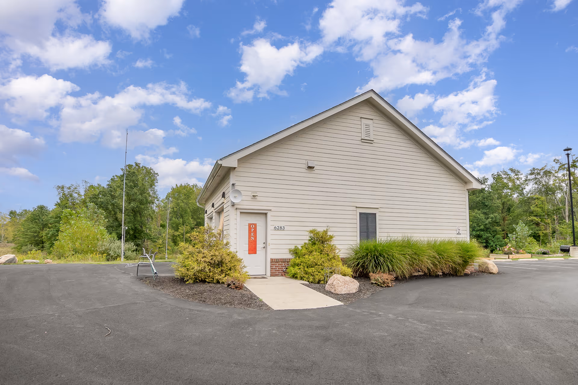 Exterior view of a small beige building with a gable roof, surrounded by greenery and a paved driveway under a partly cloudy sky. The building has a door with a red 'OPEN' sign and the number 6283 displayed next to it.