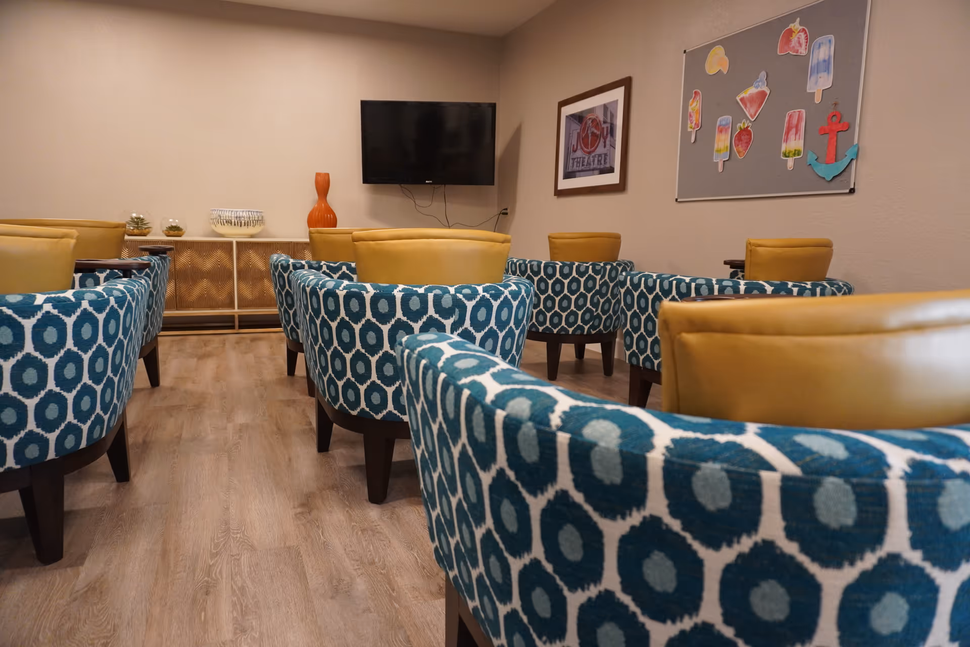 A small room with multiple blue and white patterned armchairs and yellow chairs arranged facing a wall-mounted flat screen TV. The room has light wood flooring, a sideboard with decorative items, a framed picture on the wall, and a bulletin board with colorful paper cutouts.