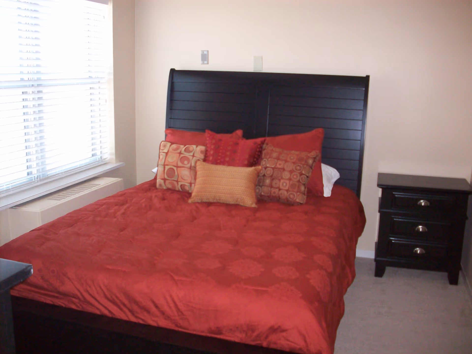 A bedroom with a large bed featuring a dark wooden headboard and a red bedspread. The bed is decorated with multiple pillows in red and orange tones. To the right of the bed is a dark wooden nightstand with three drawers. There is a window with white blinds on the left side of the room.