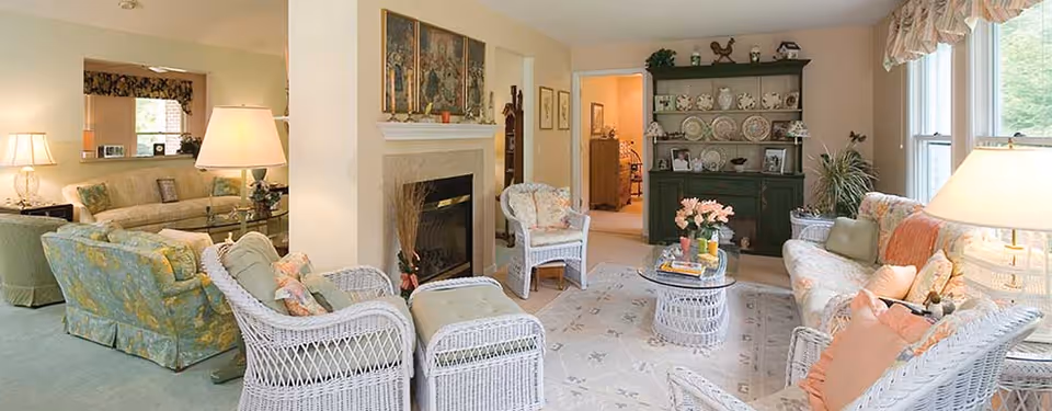 Light-filled living room with white wicker chairs and ottomans, floral sofas, a fireplace, and a green china cabinet.