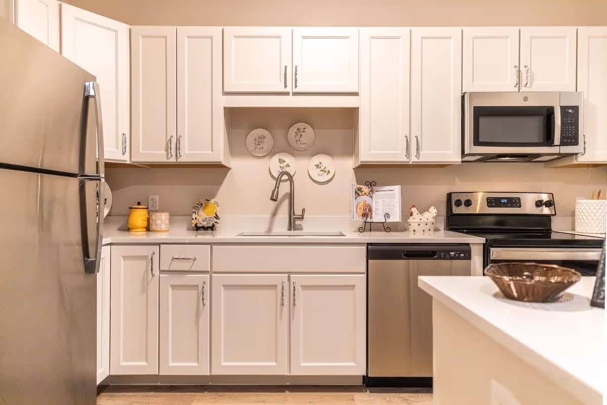 A modern kitchen with white cabinets, a stainless steel refrigerator, dishwasher, microwave, and stove. The countertop is white with a sink and faucet in the center. Decorative plates are mounted on the wall above the sink, and various kitchen items are placed on the counters.