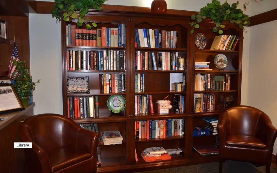 Cozy library corner with a large wooden bookshelf filled with books and decorative items, flanked by two brown leather armchairs. There are plants on top of the bookshelf and an American flag on the left side.