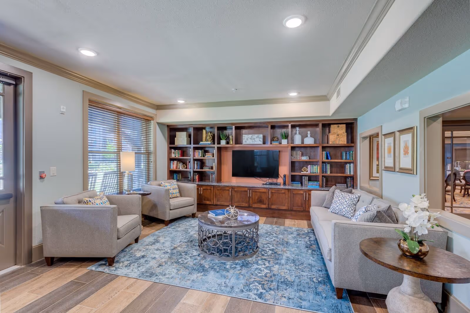 A cozy living room area with a large wooden built-in bookshelf and TV unit, two armchairs, a sofa with patterned pillows, a round coffee table on a blue patterned rug, and a side table with a white orchid plant. The room has wooden flooring, large windows with blinds, and recessed ceiling lights.