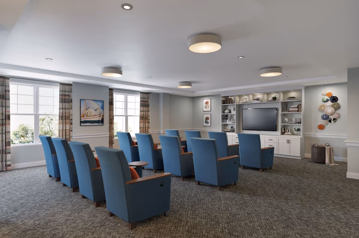 Bright common living room with rows of blue armchairs facing a large wall-mounted TV and built-in shelving.