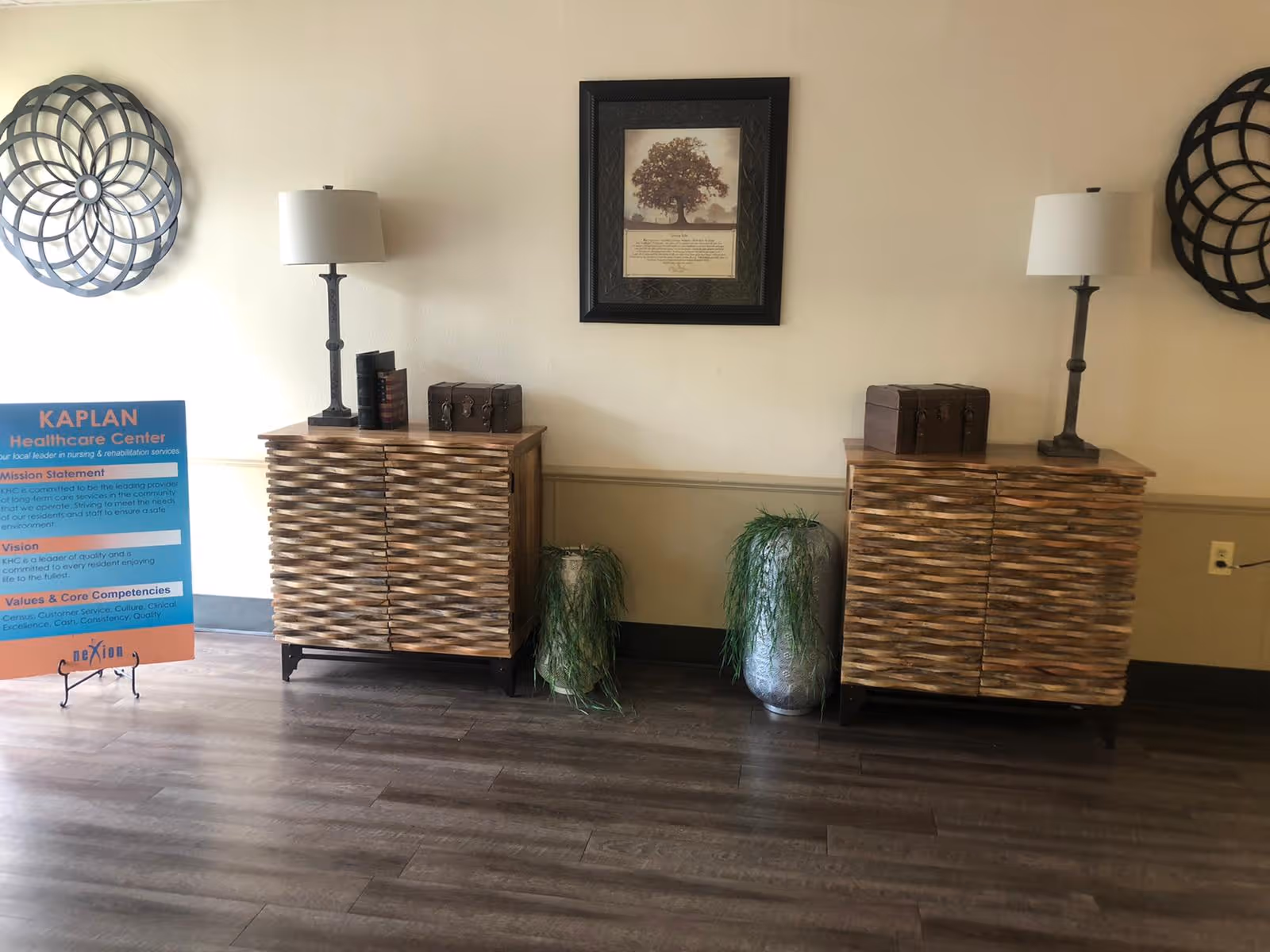 Interior view of a room at Kaplan Healthcare Center featuring two wooden cabinets with textured fronts, each topped with a table lamp and a decorative chest. Between the cabinets are two tall silver vases with green plants. A framed picture of a tree hangs on the wall above the center. On the left side, there is a blue and orange sign with the Kaplan Healthcare Center's mission statement, vision, and core competencies. The floor is wooden, and the walls are light-colored with a darker baseboard.