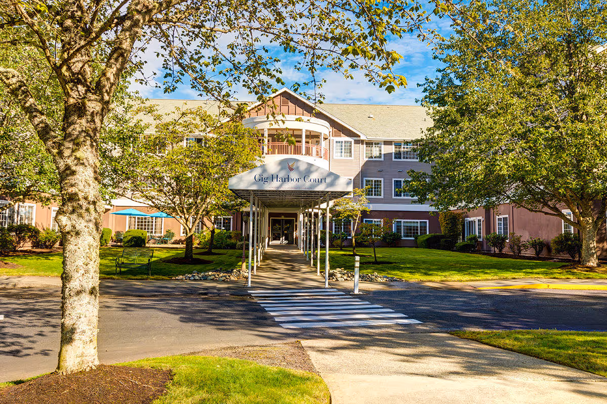 Front exterior view of Gig Harbor Court senior living facility with a covered walkway entrance, surrounded by green trees and well-maintained landscaping under a blue sky.