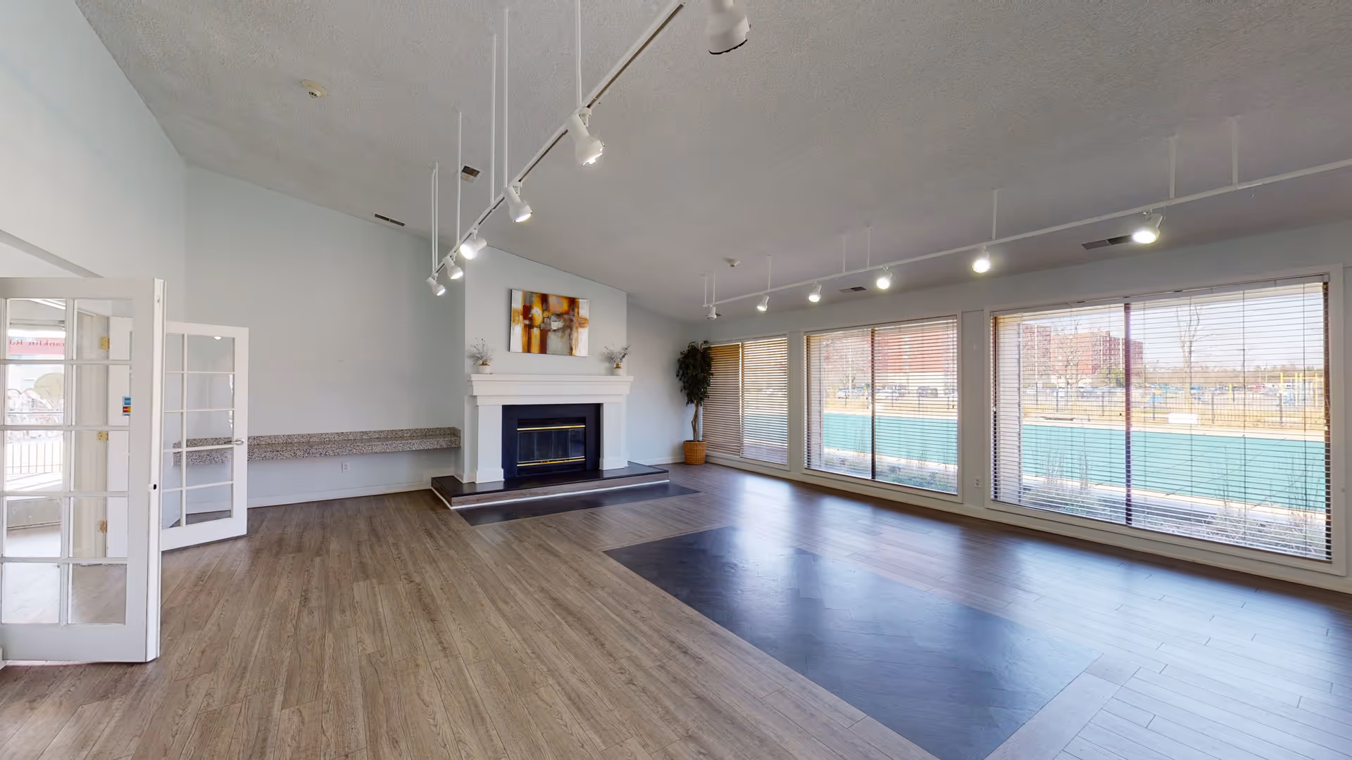 A spacious, empty common room with large windows covered by blinds letting in natural light. The room features a white fireplace with a colorful abstract painting above it, wood flooring, and track lighting on the ceiling. There is a potted plant in the corner and a granite countertop along one wall near a set of glass double doors.