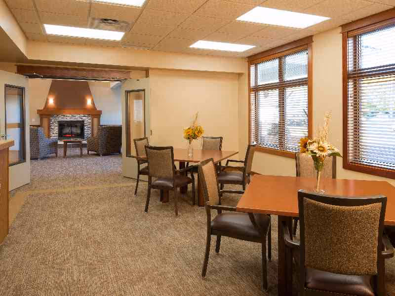Communal dining area with multiple tables and chairs, floral centerpieces, large windows, and a seating area with a fireplace in the background.
