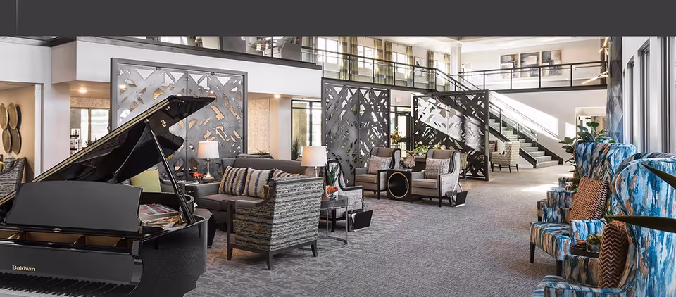 Spacious and well-lit senior living facility common area with a black grand piano on the left, multiple seating arrangements including sofas and armchairs, decorative room dividers, and a staircase leading to an upper level. The space features large windows allowing natural light and modern decor.