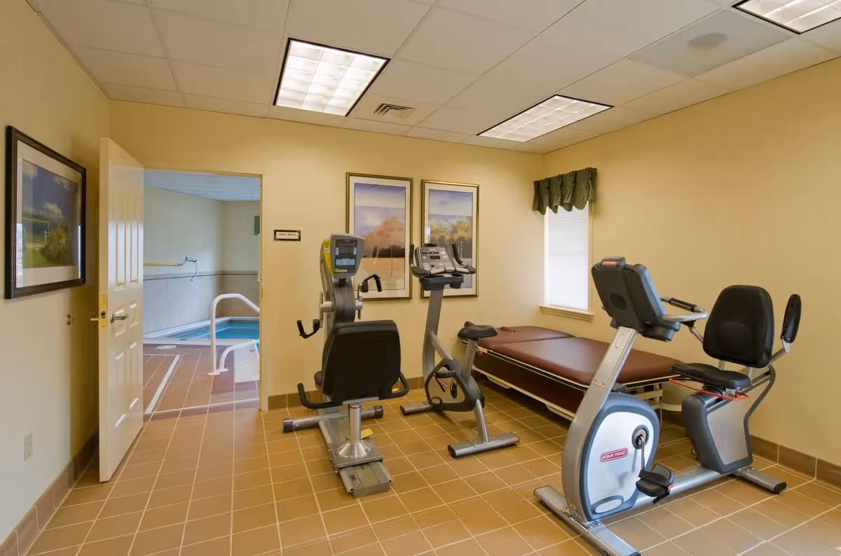 A small exercise room with two stationary recumbent bikes and a padded exercise bench. The room has beige walls, tiled floor, and two framed landscape paintings on the wall. A window with a green valance lets in natural light. An open door reveals an adjacent indoor pool area with handrails and tiled flooring.