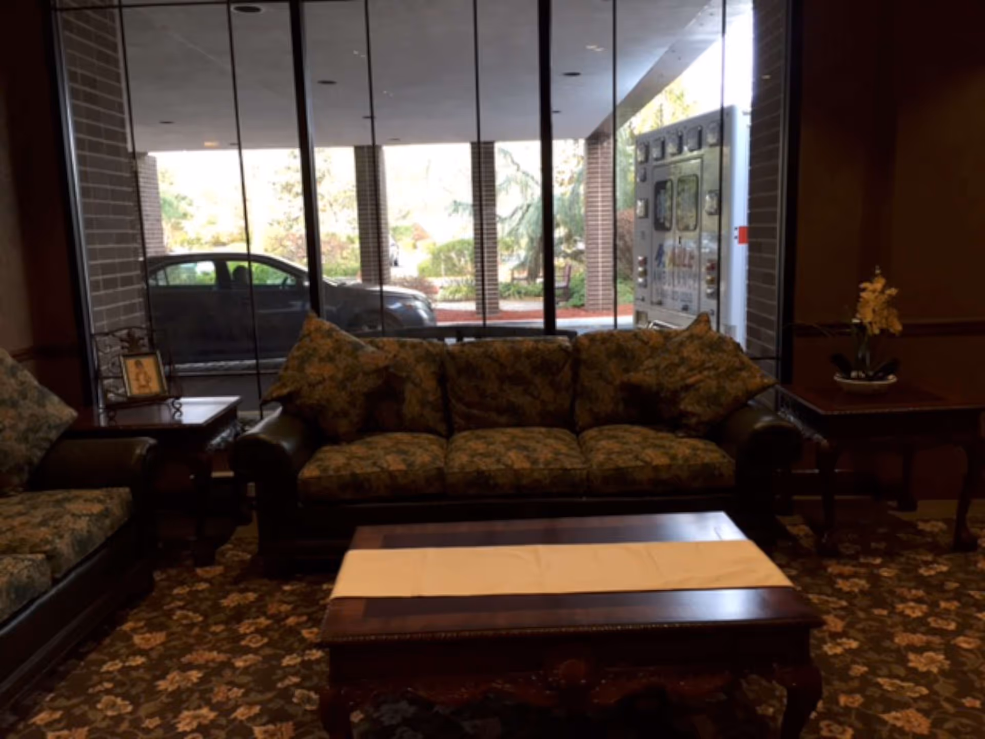 A cozy seating area inside a facility with a floral-patterned sofa and armchair, a wooden coffee table with a white runner, and side tables with decorative items. Large windows behind the sofa show a parked car and an ambulance outside.