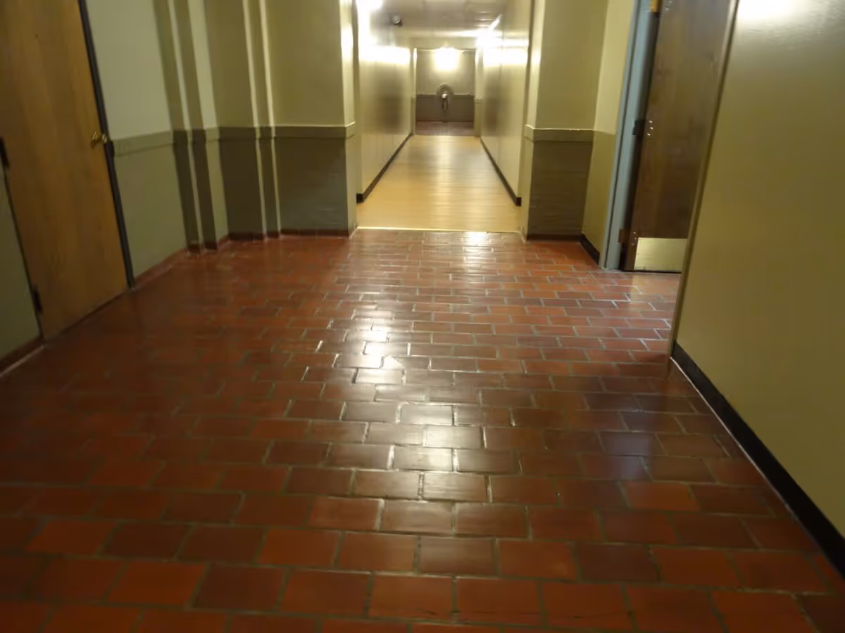 A hallway with reddish-brown tiled floor leading to a corridor with wooden flooring. The walls are painted beige with a darker beige lower half. There are wooden doors on either side of the hallway and a water fountain visible at the end of the corridor.