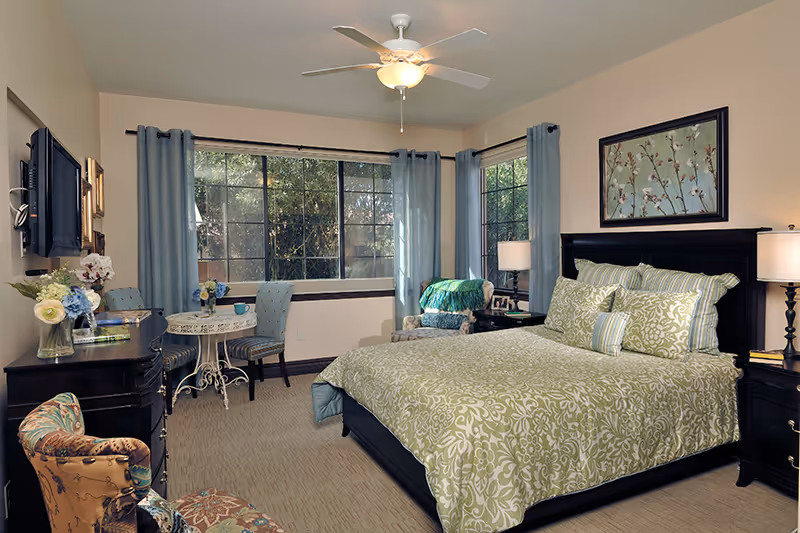 Furnished bedroom with a large bed in green patterned bedding, nightstands and lamps, seating area and small table by large windows with blue curtains.