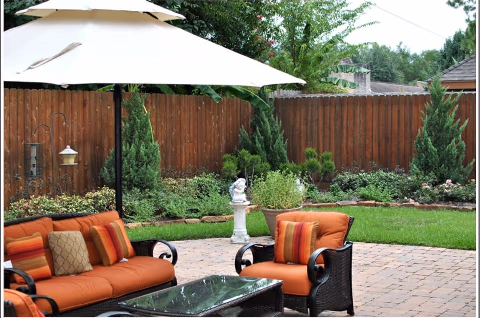 Outdoor patio area with orange cushioned wicker furniture including a sofa and chair, a glass-top coffee table, a large white umbrella, and a wooden fence with green shrubs and plants in the background.