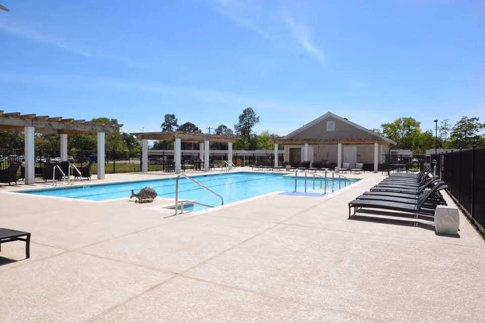 Outdoor swimming pool area with lounge chairs lined up on the right side, pergolas with seating areas on the left, and a small building in the background under a clear blue sky.