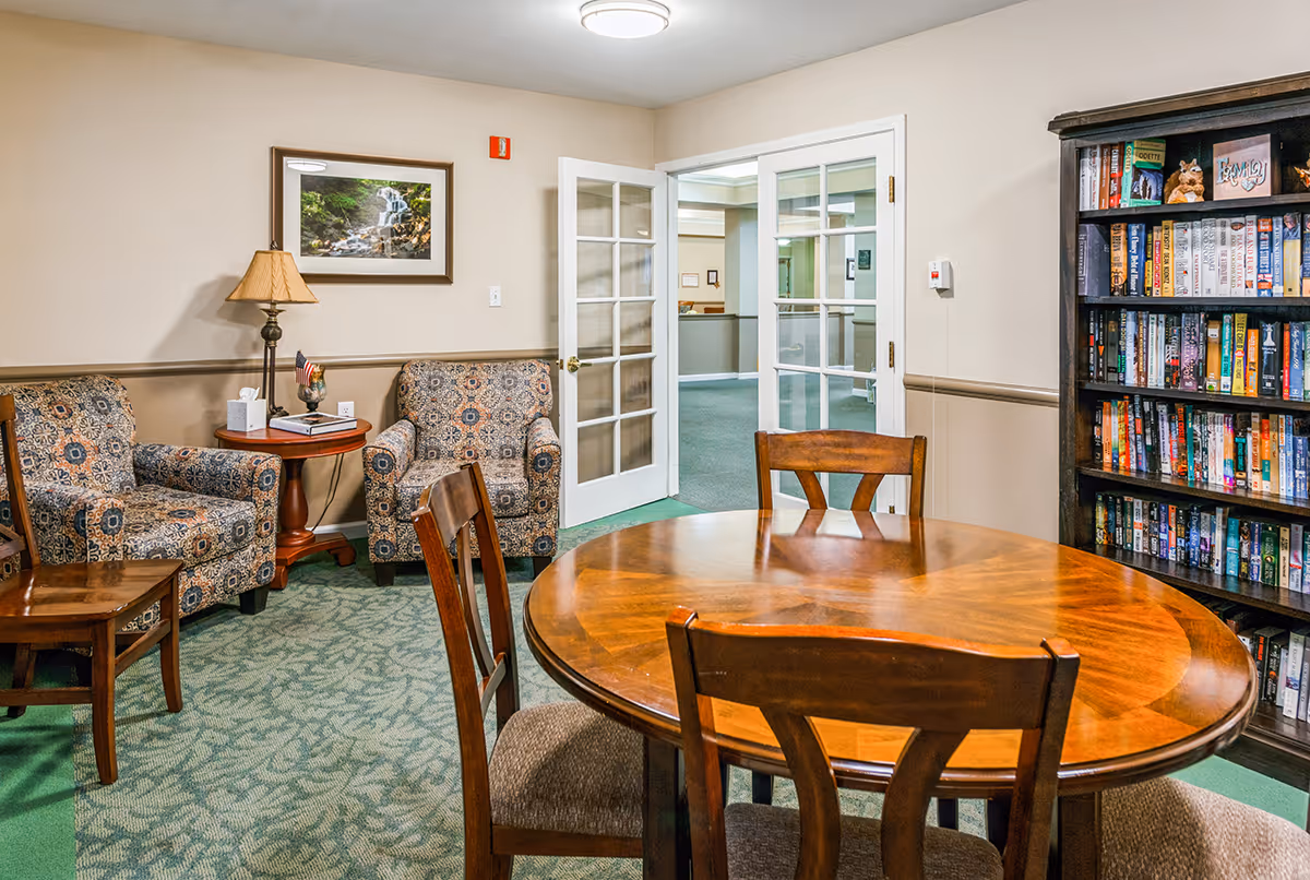 A cozy interior room with a round wooden table and four chairs in the foreground. To the left, there are two patterned armchairs with a small round wooden side table between them, holding a lamp, a tissue box, and some books. On the right side, there is a tall bookshelf filled with books and decorative items. The room has beige walls, green patterned carpet, and a framed picture of a waterfall on the wall. Double glass doors are open, showing a hallway beyond.
