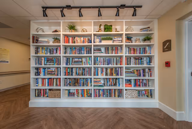 A large white bookshelf filled with numerous books and decorative items such as plants and small sculptures, set against a beige wall with wood-patterned flooring in front.
