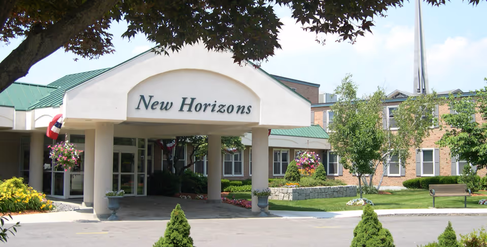 Exterior front entrance of the New Horizons senior living facility with a covered driveway, green roof, hanging flower baskets, and landscaped garden with trees and shrubs.