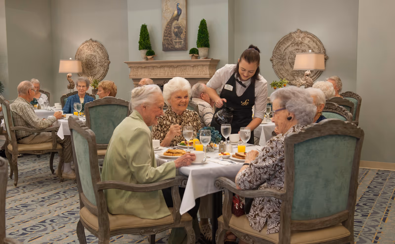 A group of elderly people sitting at tables in a dining room, enjoying a meal together. A waitress is pouring coffee for one of the women. The room is decorated with elegant chairs, a fireplace, plants, and wall art.