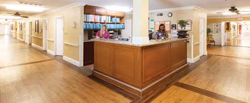 Wood reception desk in a bright senior living hallway with staff at work.
