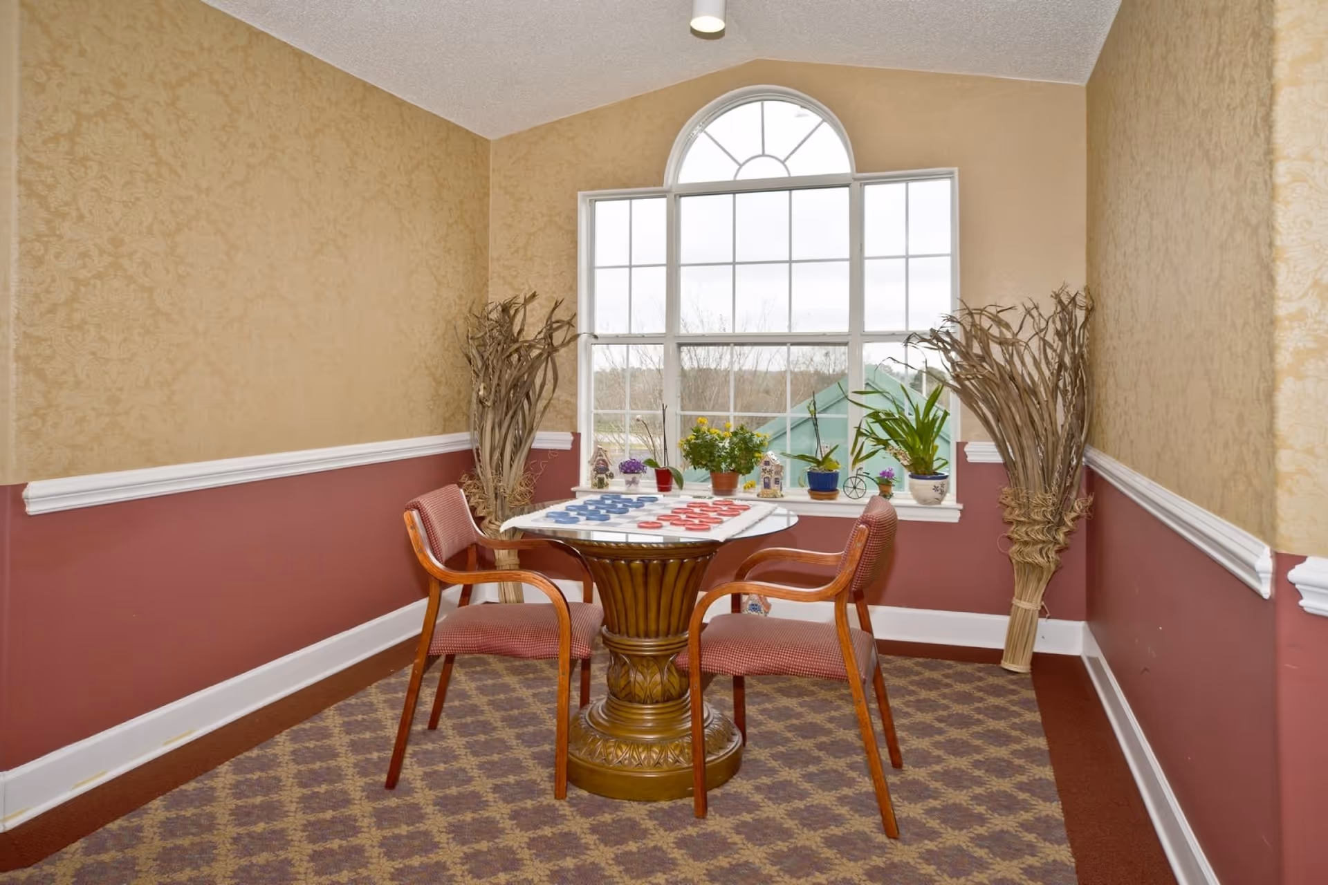A small seating area with a round wooden table and two wooden chairs with red cushions. The table has a checkerboard game set up with red and blue pieces. The room has patterned wallpaper in beige and maroon colors, a large arched window with several potted plants on the windowsill, and decorative dried branches in the corners.