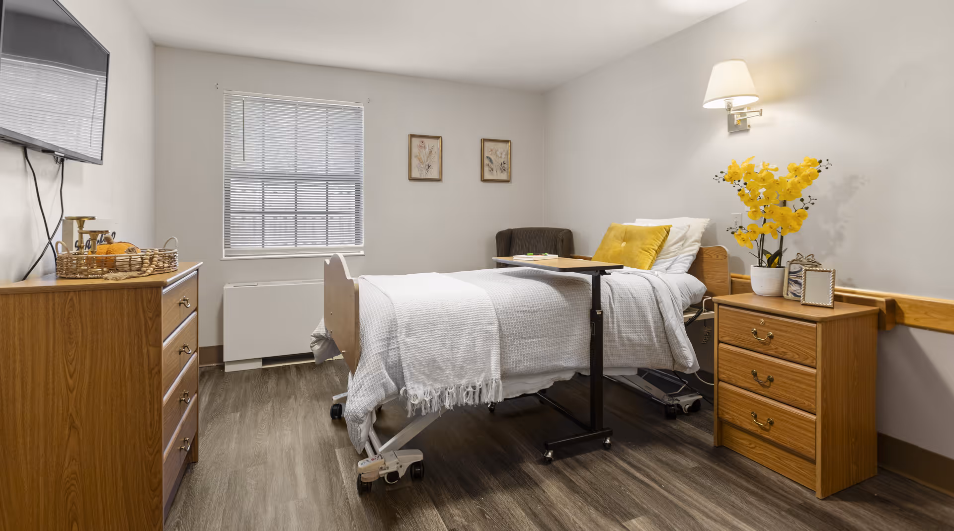 A senior living facility bedroom with a hospital-style bed covered in white bedding and a yellow pillow. There is a wooden nightstand with a lamp, a vase of yellow flowers, and a picture frame. A wooden dresser with a basket and other items is on the left side, and a flat-screen TV is mounted on the wall above it. The room has a window with blinds and two framed pictures on the wall.