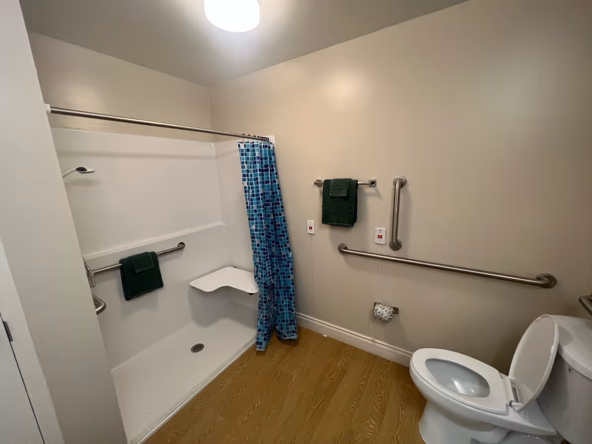A bathroom with a walk-in shower featuring a fold-down seat and grab bars, a blue patterned shower curtain, a toilet with the lid open, and green towels hanging on the wall. The floor has a wood-like finish.