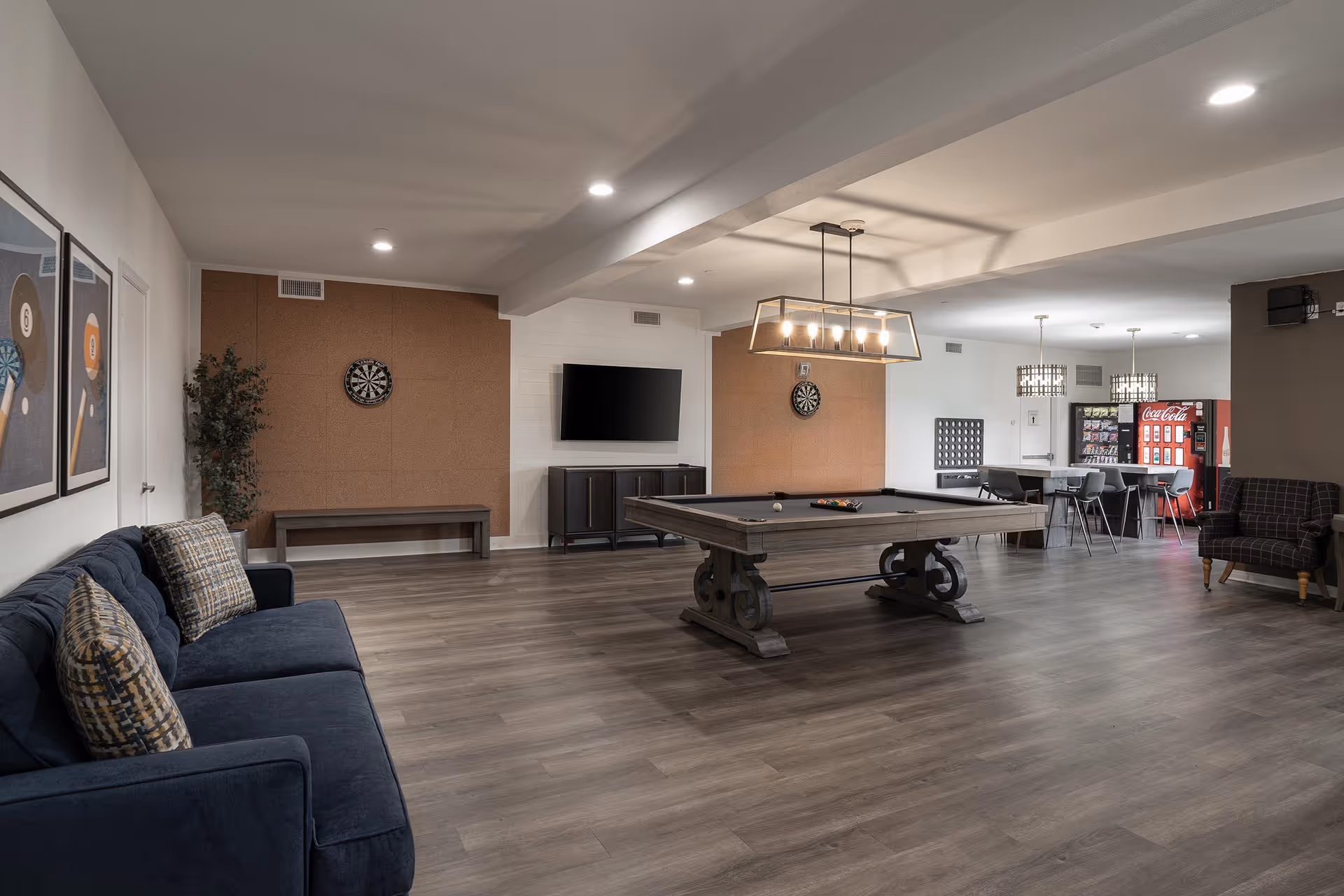 A spacious recreational room featuring a pool table with balls arranged for a game, two dartboards mounted on cork walls, a flat-screen TV on a cabinet, a blue couch with patterned pillows, a bench, a vending machine area with tables and chairs, and modern lighting fixtures.