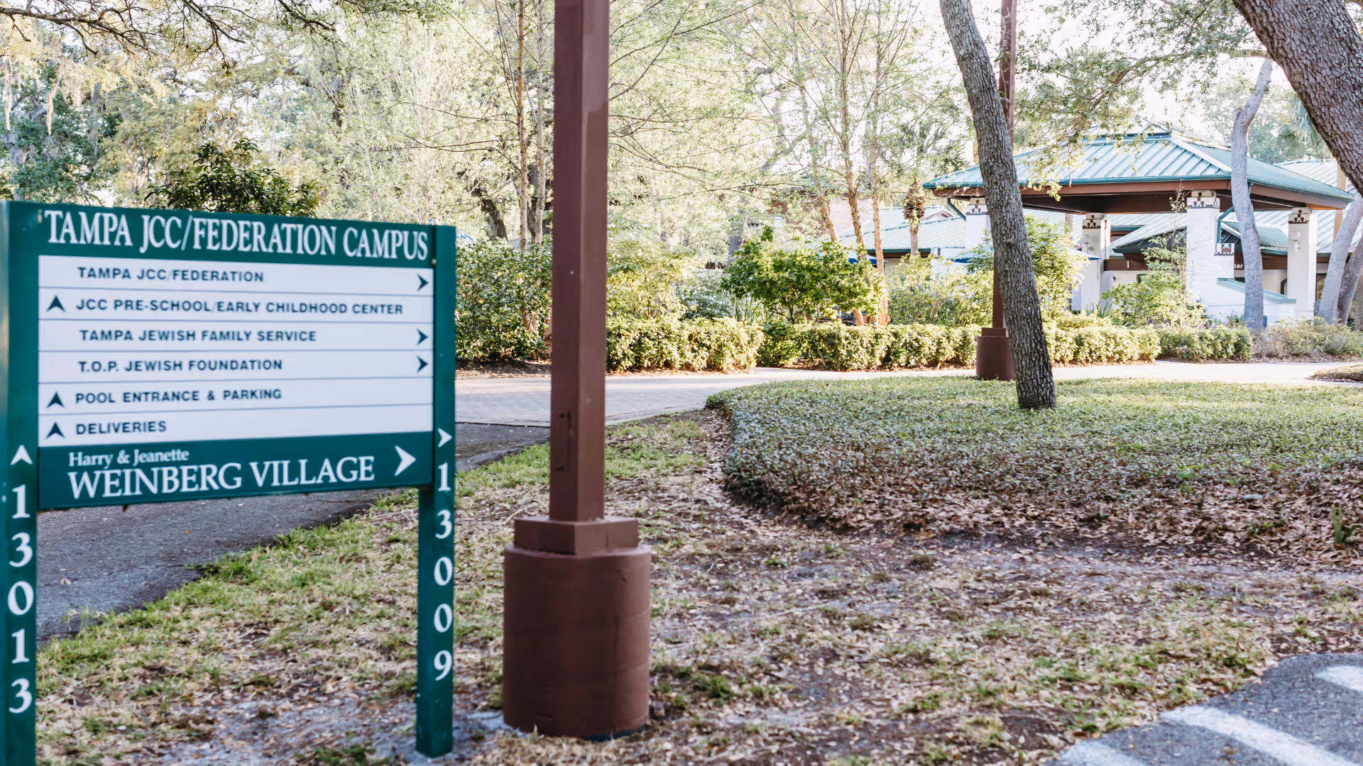 Outdoor view of the Tampa JCC/Federation Campus signpost with directions to various facilities including the Harry & Jeanette Weinberg Village. The area is surrounded by trees, bushes, and a paved pathway leading to a building with a green roof in the background.