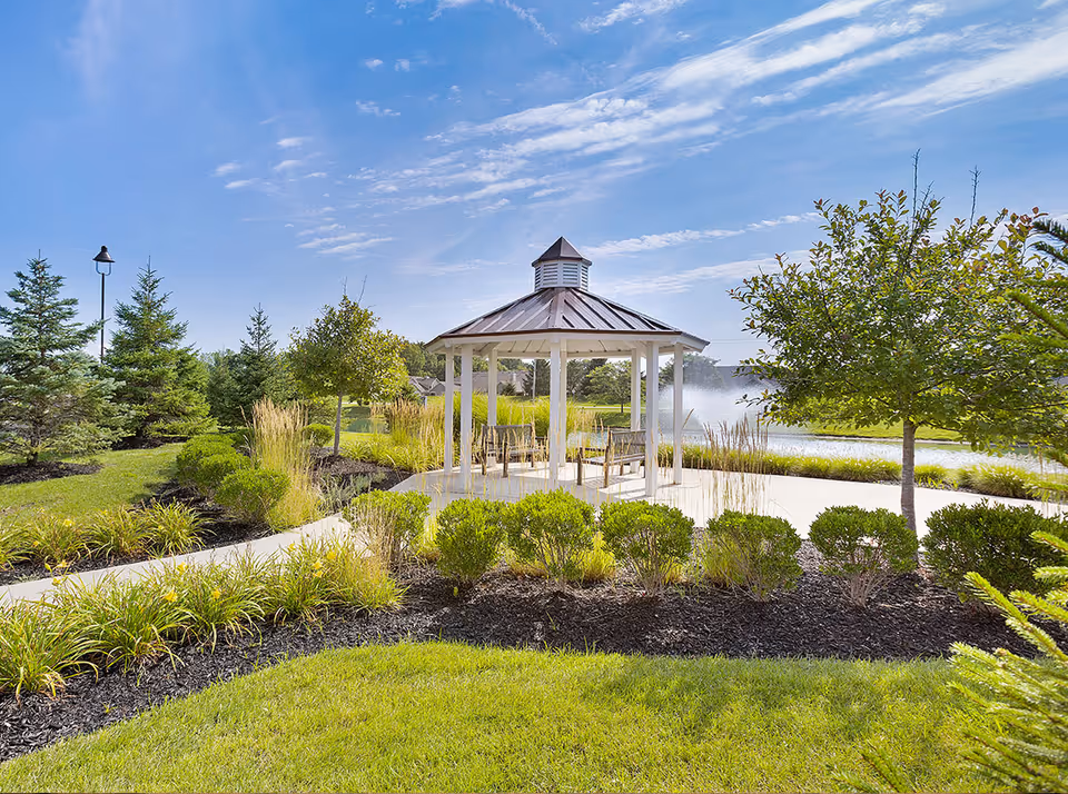 A white gazebo surrounded by landscaped gardens and a pond with a fountain under a blue sky.