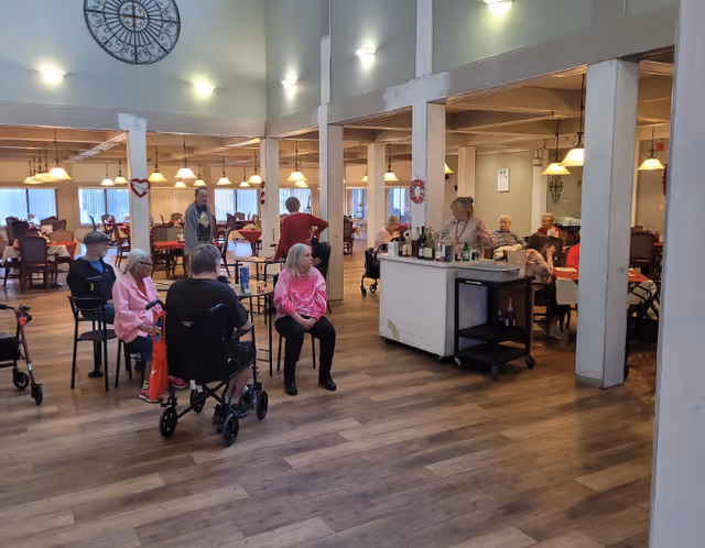 A spacious dining area in a senior living facility with several elderly residents sitting on chairs and using walkers. A woman stands behind a small bar cart with various bottles. The room has wooden floors, white pillars, and warm lighting with hanging lamps and wall sconces. A large decorative clock is mounted on the wall.