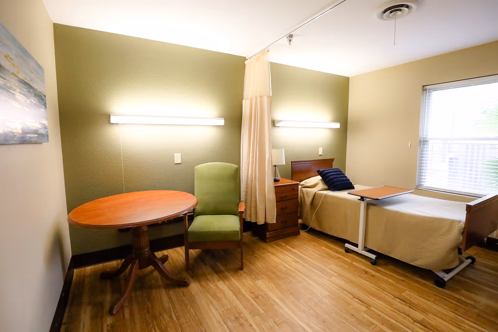 A simple, clean bedroom in a rehabilitation and healthcare center featuring a single bed with beige bedding and a dark blue pillow, a wooden bedside table with a lamp, a green upholstered chair, and a round wooden table. The room has light green and beige walls, a window with blinds, and wooden flooring.