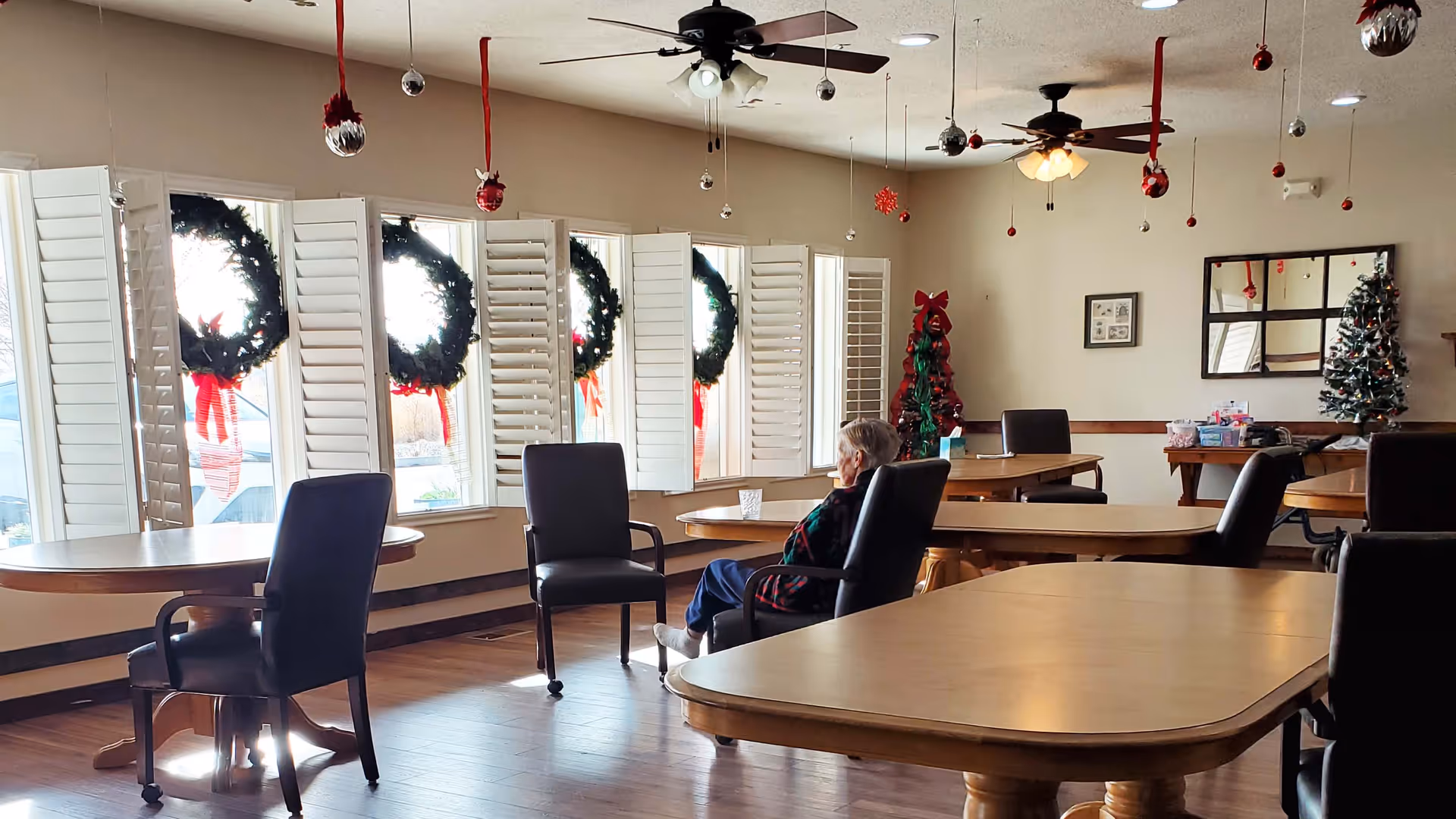 Community dining/lounge room with multiple tables and chairs, holiday wreaths on the windows, ceiling fans, and a person seated near the center.