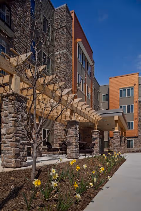Exterior view of a multi-story senior living facility with stone and wood paneling. The building features a covered patio area with seating and a wooden pergola. In the foreground, there is a landscaped garden bed with blooming yellow and white flowers along a concrete walkway under a clear blue sky.