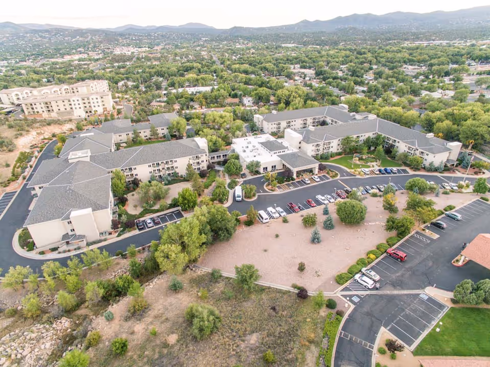Aerial view of Las Fuentes Resort Village, showing a large multi-wing building surrounded by trees and parking lots with several cars. The facility is situated in a green suburban area with hills in the background.