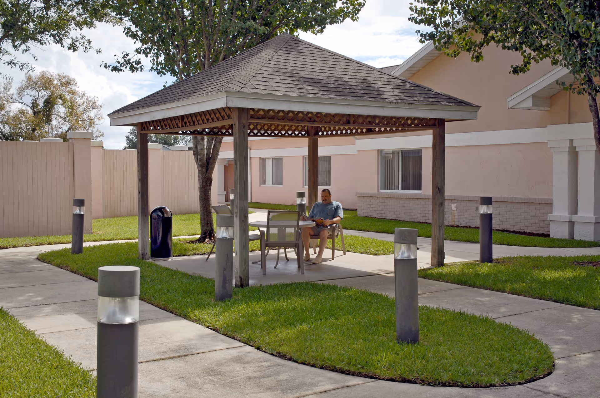 A man sitting under a wooden gazebo with a shingled roof in an outdoor courtyard area. The gazebo is surrounded by green grass and paved walkways with modern outdoor lights. In the background, there is a beige building with windows and a wooden fence.