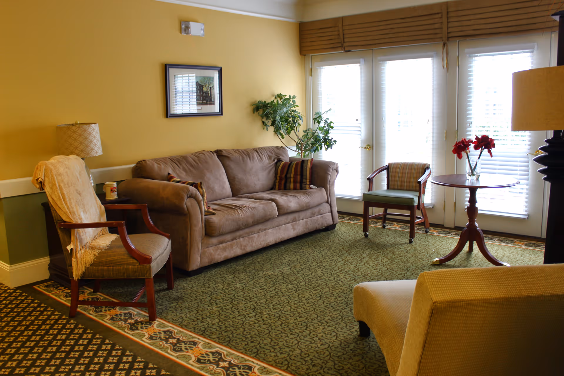 A cozy living room with a brown sofa, two armchairs, a small round wooden table with a vase of red flowers, a floor lamp, and a framed picture on a yellow wall. The room has green patterned carpet and large windows with white blinds letting in natural light.