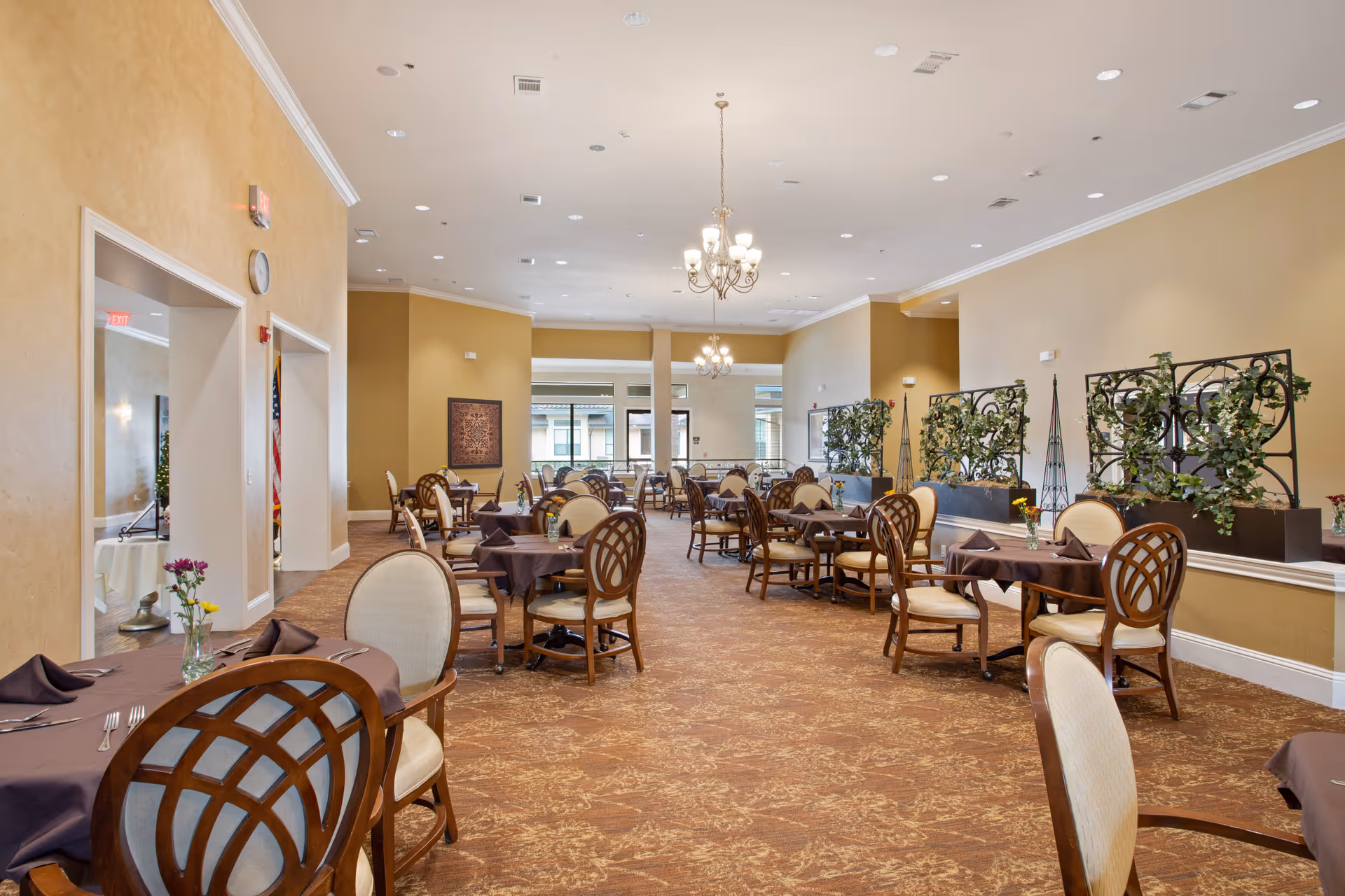 A spacious dining room with multiple round tables covered with brown tablecloths, each set with napkins and small flower vases. The room has beige walls, carpeted floors, chandeliers hanging from the ceiling, and decorative plants along one side.