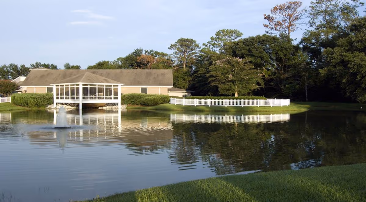 A serene outdoor scene at Live Oak Village featuring a pond with a small fountain in the foreground, a building with a screened porch extending over the water, surrounded by green grass, trees, and a white picket fence.