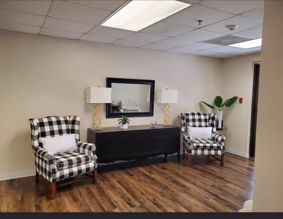 Two black-and-white checkered armchairs flank a black console table with lamps, a mirror, and a plant in a reception-style room with wood flooring.
