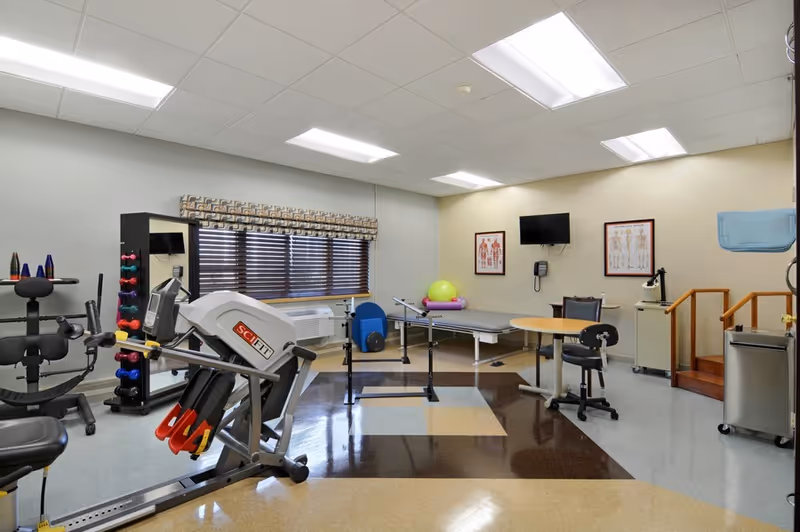 A physical therapy room with exercise equipment including a SciFit machine, dumbbells on a rack, therapy balls, a treatment table, a small round table with chairs, anatomical charts on the wall, a mounted TV, and stairs with handrails.